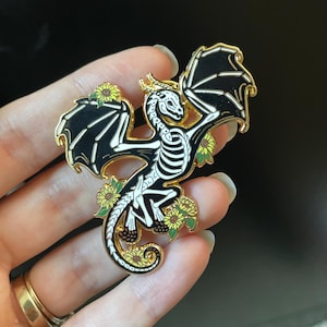 Puede incluir: Un pin de esmalte negro y blanco de un drag&oacute;n esqueleto con detalles dorados. El drag&oacute;n est&aacute; volando con las alas extendidas y tiene un dise&ntilde;o floral con girasoles amarillos.