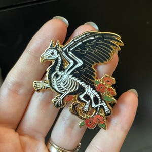 Puede incluir: Un pin de esmalte dorado con un grifo esquel&eacute;tico con alas negras y flores rojas. El grifo est&aacute; representado en un estilo anat&oacute;mico detallado.