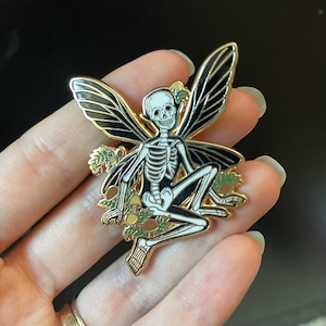 Puede incluir: Un pin de esmalte dorado con un hada esqueleto con alas negras y un cuerpo blanco. El hada est&aacute; sentada en una cama de hojas verdes y setas amarillas.