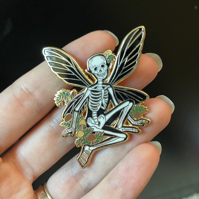 Puede incluir: Un pin de esmalte dorado con un hada esqueleto con alas negras y un cuerpo blanco. El hada est&aacute; sentada en una cama de hojas verdes y setas amarillas.