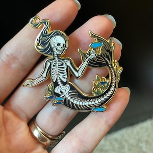 Puede incluir: Un pin de esmalte dorado con una sirena esqueleto con detalles en blanco y negro. La sirena est&aacute; rodeada de algas marinas y peque&ntilde;os peces azules.