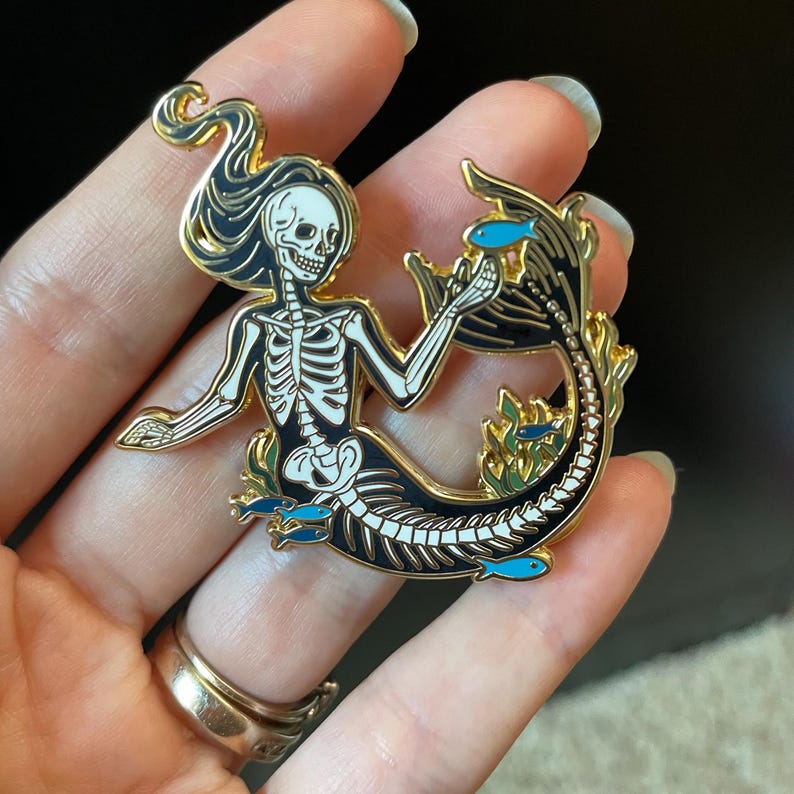 Puede incluir: Un pin de esmalte dorado con una sirena esqueleto con detalles en blanco y negro. La sirena est&aacute; rodeada de algas marinas y peque&ntilde;os peces azules.