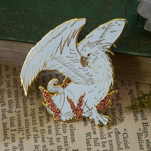 May include: A gold-toned enamel pin depicting a white griffin with red eyes and red flowers. The griffin's wings are spread and its head is turned to the side. The pin is a detailed and intricate piece of artwork.