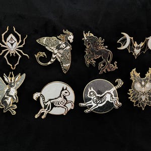 May include: A collection of enamel pins featuring various fantasy creatures. Designs include a spider, moth, unicorn, bat, rabbit, and cat skeletons. The pins have gold and silver accents with black and white details, set against a black background.