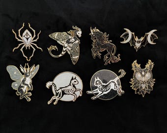 Pode incluir: Uma coleção de alfinetes de esmalte com várias criaturas de fantasia. Os designs incluem uma aranha, uma mariposa, um unicórnio, um morcego, um coelho e esqueletos de gato. Os alfinetes têm detalhes dourados e prateados com detalhes em preto e branco.