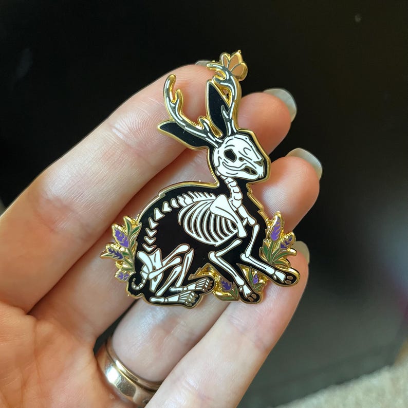 Puede incluir: Un pin de esmalte dorado con un conejo esquel&eacute;tico negro con cuernos y flores moradas. El conejo est&aacute; sentado sobre sus patas traseras con las patas delanteras extendidas.