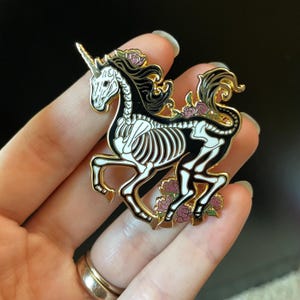 Puede incluir: Pin de esmalte dorado con un unicornio esqueleto negro y blanco con flores rosas. El unicornio corre con el cuerno apuntando hacia adelante.