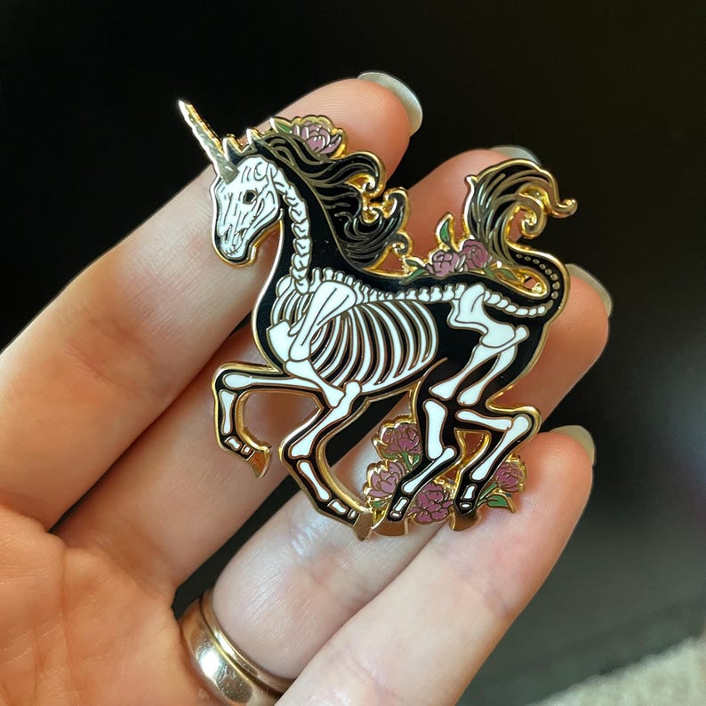 Puede incluir: Pin de esmalte dorado con un unicornio esqueleto negro y blanco con flores rosas. El unicornio corre con el cuerno apuntando hacia adelante.
