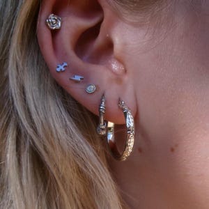 May include: Close-up of an ear adorned with multiple silver earrings. The ear features a rose stud, puzzle piece stud, lightning bolt stud, and a small round stud. Two patterned hoop earrings complete the look.