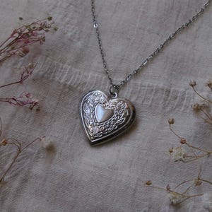May include: A silver-toned heart-shaped locket pendant suspended from a matching delicate link chain. The locket features an intricate embossed floral pattern surrounding a smooth, central heart-shaped inlay. The metal has a polished finish.