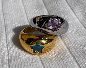 Pode incluir: Dois anéis em forma de cúpula. Um é prateado com uma gema rosa em forma de estrela, o outro é dourado com uma gema azul em forma de estrela. Ambos os anéis estão sobre uma superfície branca texturizada.