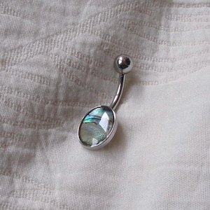 May include: A curved barbell belly button ring featuring a polished silver-tone metal finish and a single oval-shaped inlay of iridescent abalone shell. The jewellery piece has a small spherical bead at the top and a larger oval setting at the bottom.