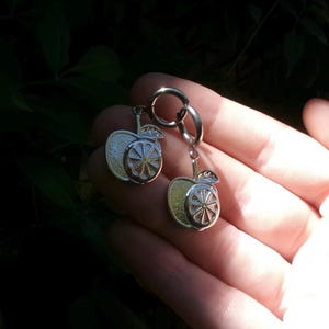 Puede incluir: Pendientes plateados con forma de manzana y diseño de rodaja de cítricos. Los pendientes se sostienen en una mano sobre un fondo de follaje verde. Los colgantes en forma de manzana tienen un aspecto detallado y texturizado.