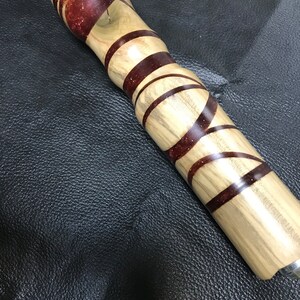 Puede incluir: Un mango cilíndrico fabricado en madera de color claro y resina de color marrón rojizo oscuro. El diseño presenta bandas alternas de madera y resina con patrones en espiral y partículas de purpurina incrustadas en las secciones oscuras.