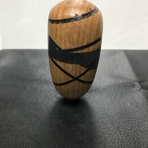 Op de afbeelding: Een glad, afgerond houten object met een lichtbruine nerf en opvallende zwarte harsinleg die kruisende lijnen en abstracte vormen op het oppervlak vormt. Het stuk heeft een gepolijste afwerking en een taps toelopende, eivormige cilindrische vorm.