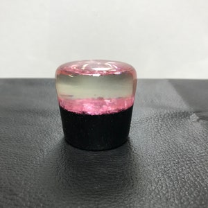 May include: A cylindrical knob featuring a solid black base and a translucent top section. The upper portion contains a layer of pink glitter embedded within clear resin, creating a distinct contrast between the opaque black bottom and the clear pink top.