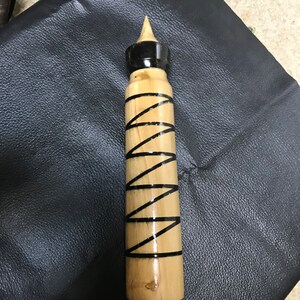 Può includere: Un utensile cilindrico in legno con finitura color marrone chiaro, caratterizzato da un motivo a spirale nera che avvolge il corpo. La parte superiore presenta una punta in legno sopra una fascia nera, mentre la base ha un piccolo componente.