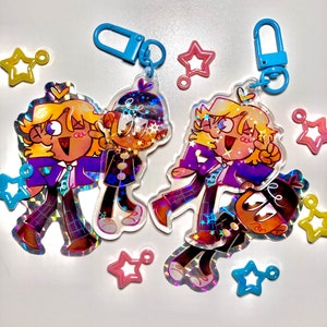 May include: Two clear acrylic keychains with cartoon characters in purple and black outfits, with holographic details. Each keychain has a blue clasp. Star-shaped charms in pink, yellow, and blue are scattered around.