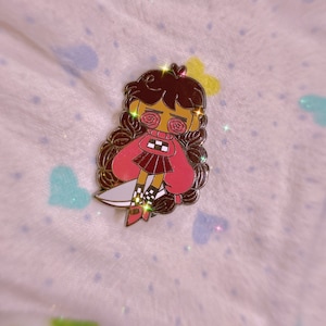 以下が含まれることがあります： 茶色の髪、赤い服、ナイフを持ったキャラクターが描かれた、漫画スタイルのエナメルピン。 ピンは金の輪郭で、柔らかい模様の背景に配置されています。 キャラクターの目はスパイラルデザインです。