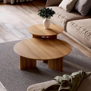 Puede incluir: Un conjunto de dos mesas de centro redondas de madera con un acabado de madera natural. Las mesas están anidadas, con la mesa más pequeña sentada encima de la mesa más grande. Las mesas tienen un diseño simple y moderno y serían una gran adición a cualquier sala de estar.