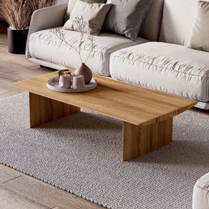 Peut inclure: Un salon moderne avec un canapé clair orné de coussins, une table basse rectangulaire en bois et un tapis gris. La table présente un plateau, des bougies et un vase avec une plante. L'ambiance est neutre et apaisante.
