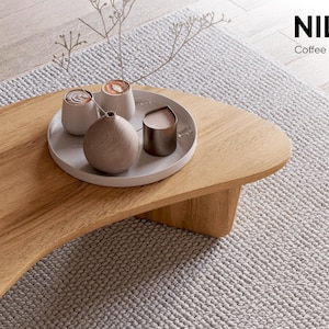 May include: A light oak coffee table with a curved edge. A white tray holds small cups, a vase, and a metal container. The table sits on a light grey textured rug. The text "NILS Coffee table" is in the upper right corner.