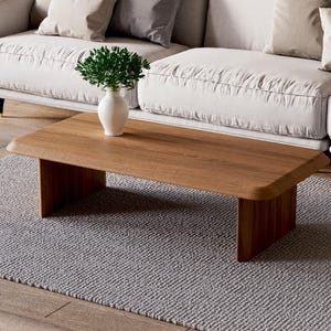 K&ouml;nnte beinhalten: Ein hellbrauner Holz-Couchtisch mit einer rechteckigen Tischplatte und zwei dicken, rechteckigen Beinen. Der Tisch steht auf einem grauen Teppich vor einem wei&szlig;en Sofa.