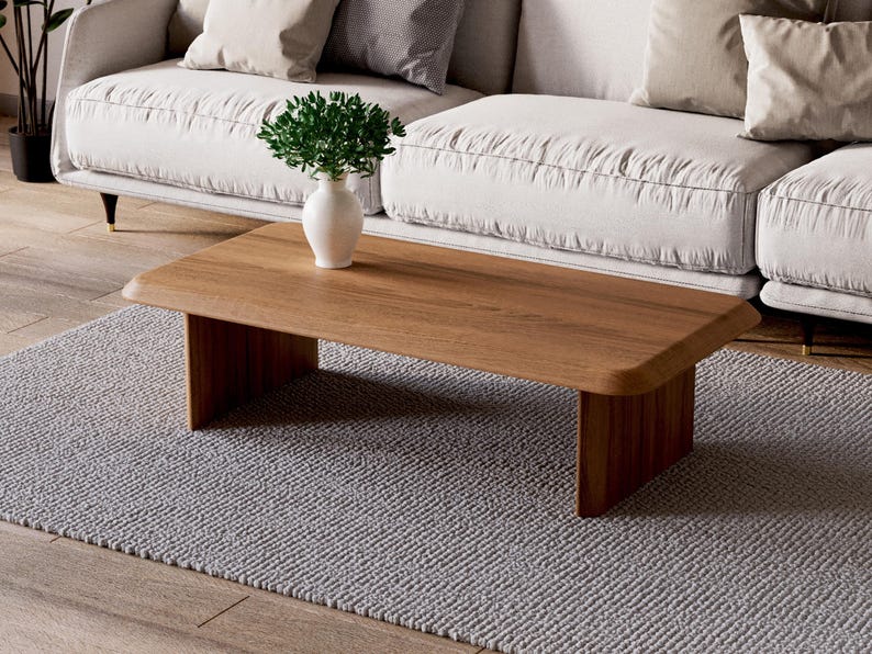 K&ouml;nnte beinhalten: Ein hellbrauner Holz-Couchtisch mit einer rechteckigen Tischplatte und zwei dicken, rechteckigen Beinen. Der Tisch steht auf einem grauen Teppich vor einem wei&szlig;en Sofa.