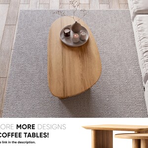Puede incluir: Una mesa de centro de madera clara con forma org&aacute;nica, colocada sobre una alfombra gris. La mesa tiene una peque&ntilde;a bandeja con velas y un jarr&oacute;n. El texto "EXPLORE MORE DESIGNS OF COFFEE TABLES!" es visible.