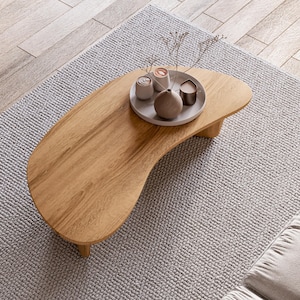 May include: A light brown, kidney-shaped wooden coffee table with a small, round tray holding decorative items. The table sits on a light grey rug, with a wooden floor in the background. The tray holds a small vase and candles.