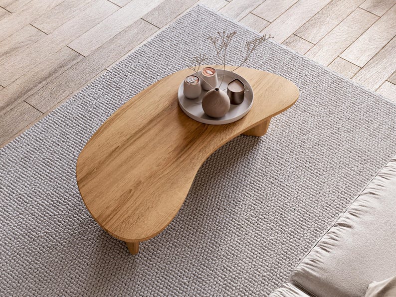 May include: A light brown, kidney-shaped wooden coffee table with a small, round tray holding decorative items. The table sits on a light grey rug, with a wooden floor in the background. The tray holds a small vase and candles.