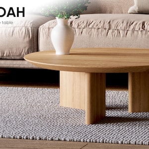 May include: A light wood coffee table with a rounded top and a solid base. A white vase with greenery sits on the table. The text "NOAH Coffee table" is in the upper left corner. The table is on a gray rug.