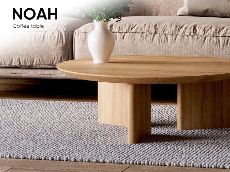 May include: A light wood coffee table with a rounded top and a solid base. A white vase with greenery sits on the table. The text "NOAH Coffee table" is in the upper left corner. The table is on a gray rug.