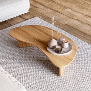 May include: A light brown, kidney-shaped wooden coffee table with a decorative tray holding small vases and candles. The table sits on a light grey rug. The table is approximately 122 cm long. A white sofa is visible in the background.