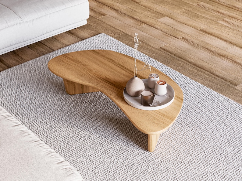 May include: A light brown, kidney-shaped wooden coffee table with a decorative tray holding small vases and candles. The table sits on a light grey rug. The table is approximately 122 cm long. A white sofa is visible in the background.