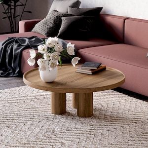 May include: A round wooden coffee table with four legs sits on a beige rug in front of a pink sofa. A vase with white flowers sits on the table, and a stack of books is next to it.