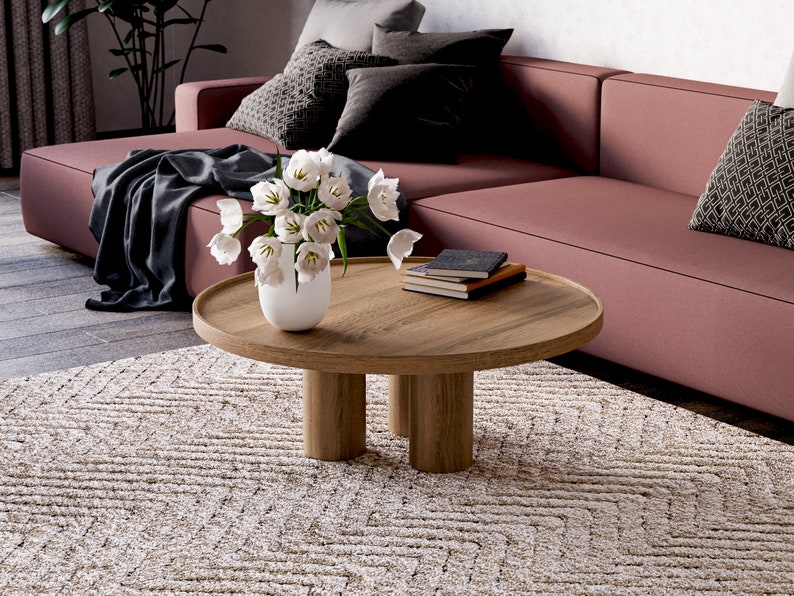 May include: A round wooden coffee table with four legs sits on a beige rug in front of a pink sofa. A vase with white flowers sits on the table, and a stack of books is next to it.