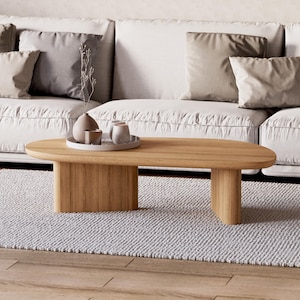 Puede incluir: Una mesa de centro ovalada de roble claro con un dise&ntilde;o minimalista. Se encuentra sobre una alfombra gris claro, frente a un sof&aacute; blanco con cojines neutros. Hay objetos decorativos en una bandeja sobre la mesa.