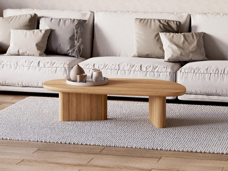 Puede incluir: Una mesa de centro ovalada de roble claro con un dise&ntilde;o minimalista. Se encuentra sobre una alfombra gris claro, frente a un sof&aacute; blanco con cojines neutros. Hay objetos decorativos en una bandeja sobre la mesa.