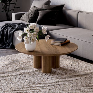 May include: A modern living room scene featuring a round wooden coffee table with three cylindrical legs. A white vase holds white tulips. A gray sectional sofa with patterned pillows and a patterned rug complete the scene.