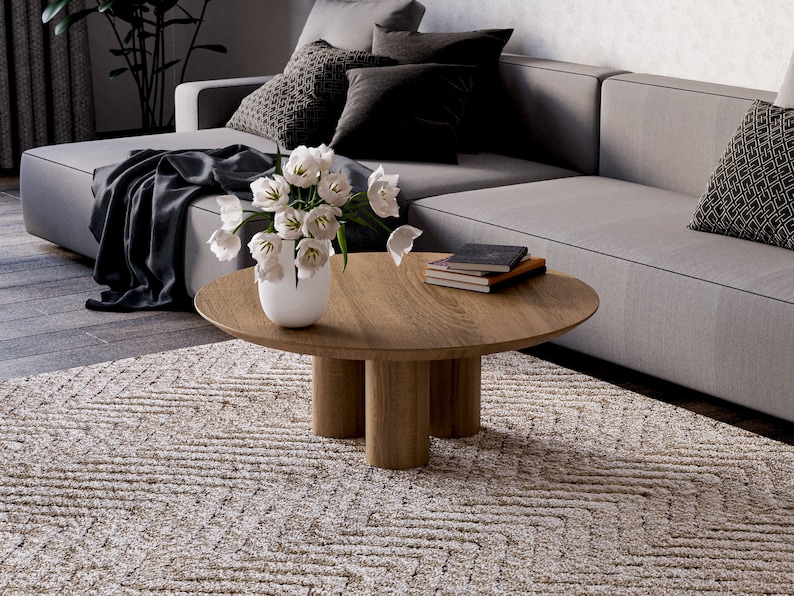 May include: A modern living room scene featuring a round wooden coffee table with three cylindrical legs. A white vase holds white tulips. A gray sectional sofa with patterned pillows and a patterned rug complete the scene.