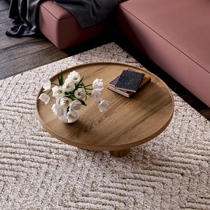 May include: A round wooden coffee table with a white vase of flowers and a stack of books on top. The table is sitting on a cream-coloured rug with a geometric pattern.