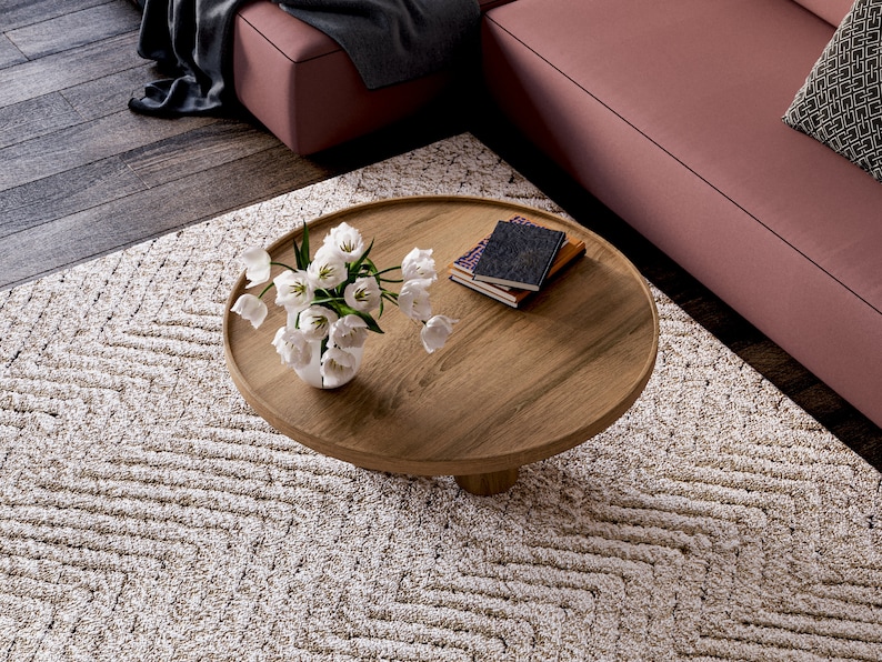 May include: A round wooden coffee table with a white vase of flowers and a stack of books on top. The table is sitting on a cream-coloured rug with a geometric pattern.