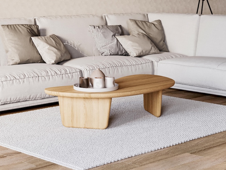 May include: A contemporary living room with a white sofa and cushions. A wooden, oval coffee table, approximately 120 cm long, rests on a white rug. Decorative items are on the table.