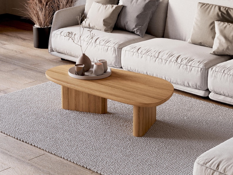 Puede incluir: Una mesa de centro ovalada de madera clara, sobre una alfombra gris clara, con un sof&aacute; blanco al fondo. La mesa tiene una veta de madera natural y un dise&ntilde;o minimalista. Objetos decorativos sobre una bandeja.