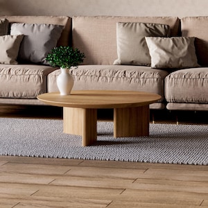 May include: A modern living room scene featuring a light brown sofa with several beige pillows. A round, light wood coffee table sits on a gray rug. A white vase with greenery is on the table. The floor is made of wood.