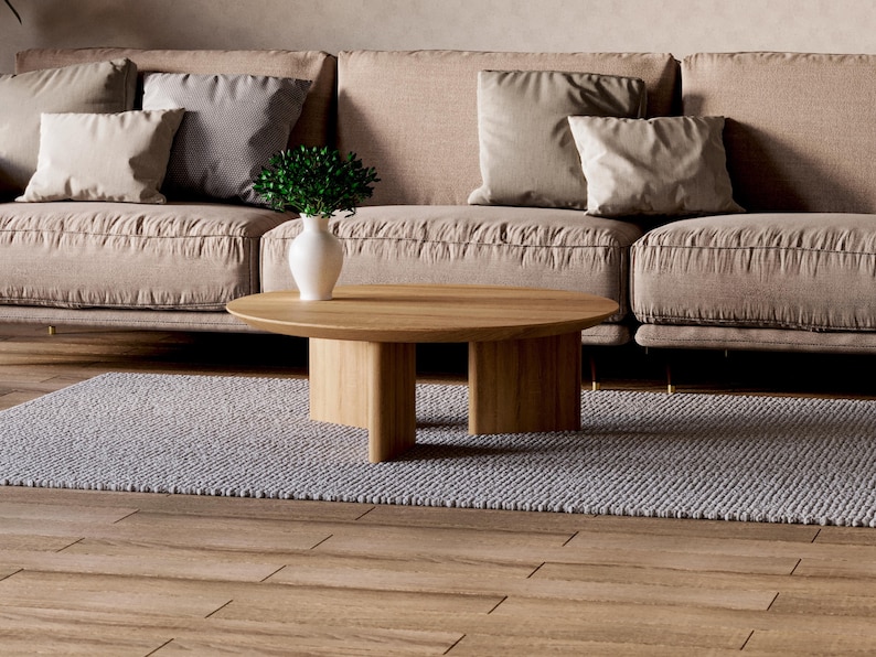 May include: A modern living room scene featuring a light brown sofa with several beige pillows. A round, light wood coffee table sits on a gray rug. A white vase with greenery is on the table. The floor is made of wood.