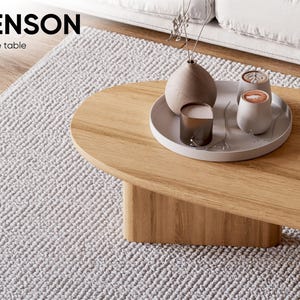 Puede incluir: Una mesa de centro de madera clara con bordes redondeados y una base s&oacute;lida. Una bandeja blanca sobre la mesa contiene un jarr&oacute;n, dos tazas peque&ntilde;as y una vela. El texto "BENSON Coffee table" est&aacute; en la esquina superior izquierda.