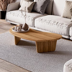 May include: A light wood, kidney-shaped coffee table with a natural finish sits on a grey rug. The table holds a tray with small white cups and a brown vase. A white sofa with beige cushions is in the background.