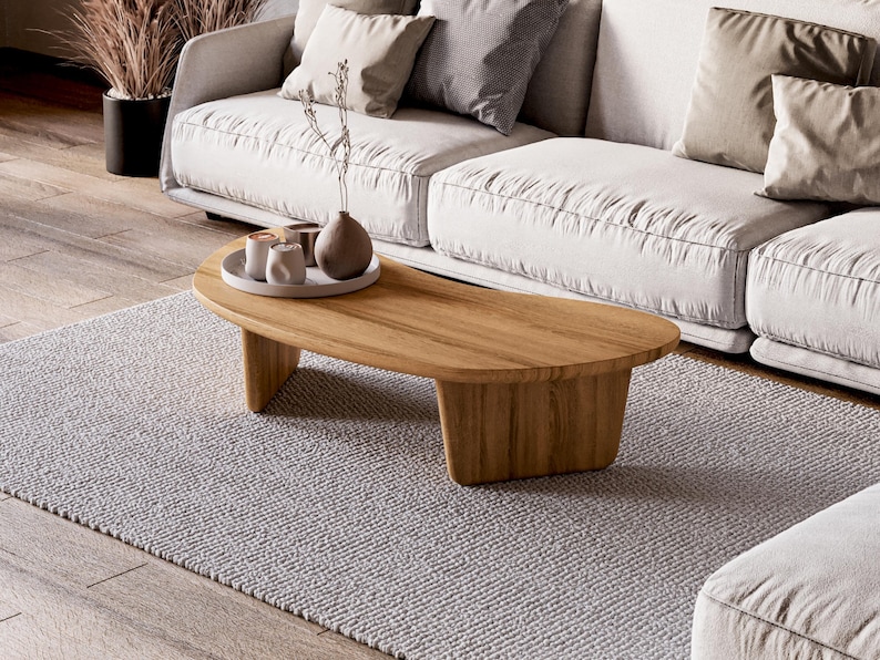 May include: A light wood, kidney-shaped coffee table with a natural finish sits on a grey rug. The table holds a tray with small white cups and a brown vase. A white sofa with beige cushions is in the background.